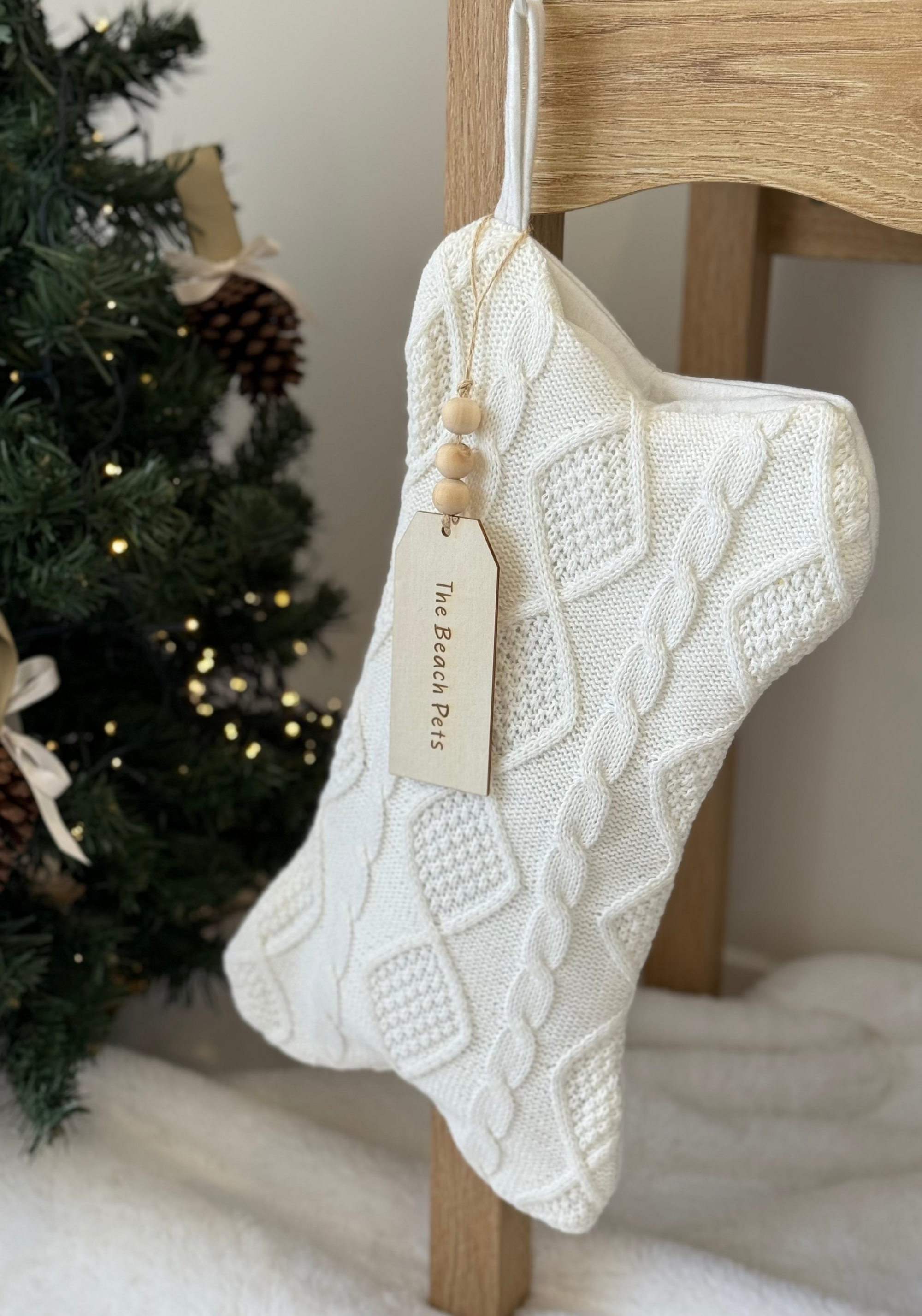 Knitted white stocking with a tag hanging on a wooden chair next to a Christmas tree.
