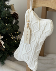 Knitted white stocking with a tag hanging on a wooden chair next to a Christmas tree.